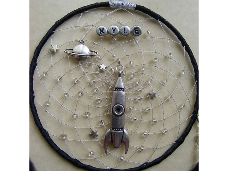 Pu&ograve; includere: Un acchiappasogni nero con una rete di perline argentate, che presenta un razzo argentato, un pianeta argentato con anelli e perline argentate che compongono il nome "KYLE".