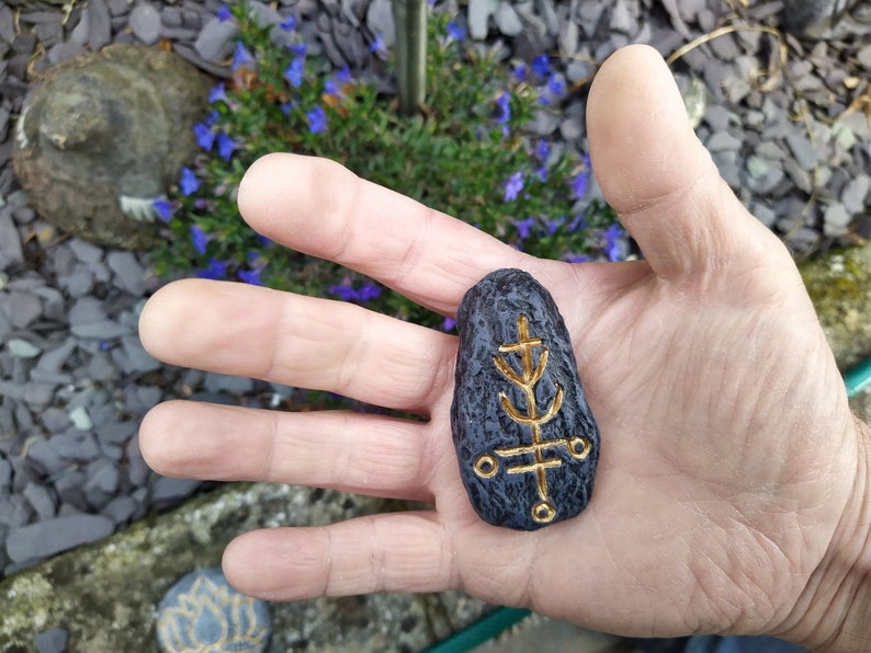 May include: A black stone with a gold tree-like symbol carved into it. The stone is being held in a hand.