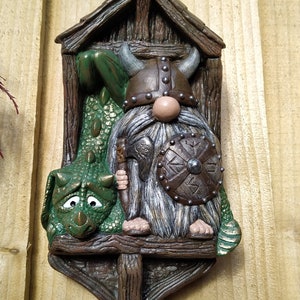 K&ouml;nnte beinhalten: Skurrile Wanddekoration mit einem Wikinger-Zwerg und einem gr&uuml;nen Drachen. Der Zwerg tr&auml;gt einen geh&ouml;rnten Helm und h&auml;lt Schild und Axt. Der Drache hat einen traurigen Gesichtsausdruck. Das St&uuml;ck besteht aus braunem, strukturiertem Material.