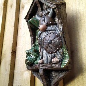 K&ouml;nnte beinhalten: Ein skurriles Wandbild mit einem Wikinger-Zwerg und einem gr&uuml;nen Drachen. Der Zwerg tr&auml;gt einen geh&ouml;rnten Helm und h&auml;lt einen Schild. Der Drache sitzt neben dem Zwerg. Das St&uuml;ck besteht aus einem braunen, strukturierten Material.