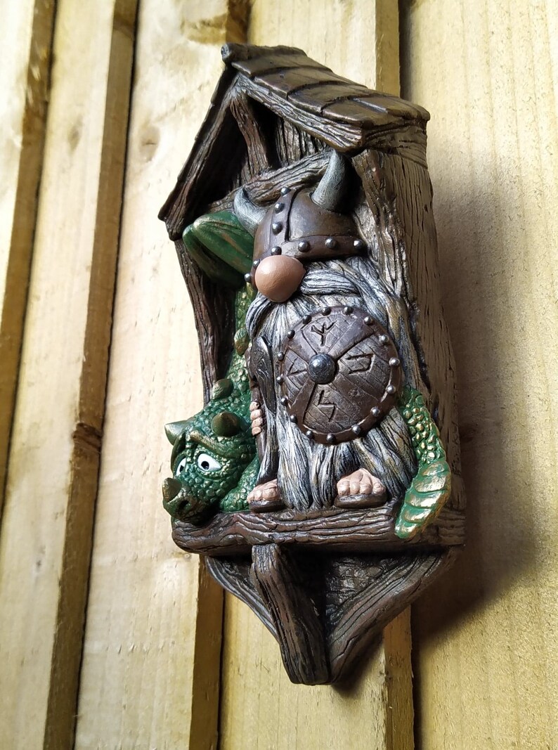 K&ouml;nnte beinhalten: Ein skurriles Wandbild mit einem Wikinger-Zwerg und einem gr&uuml;nen Drachen. Der Zwerg tr&auml;gt einen geh&ouml;rnten Helm und h&auml;lt einen Schild. Der Drache sitzt neben dem Zwerg. Das St&uuml;ck besteht aus einem braunen, strukturierten Material.