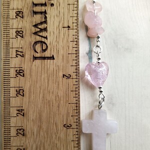 Puede incluir: Un brazalete de charms rosa y plateado con una cruz, corazones y cuentas. El brazalete mide aproximadamente 15 cm de largo.