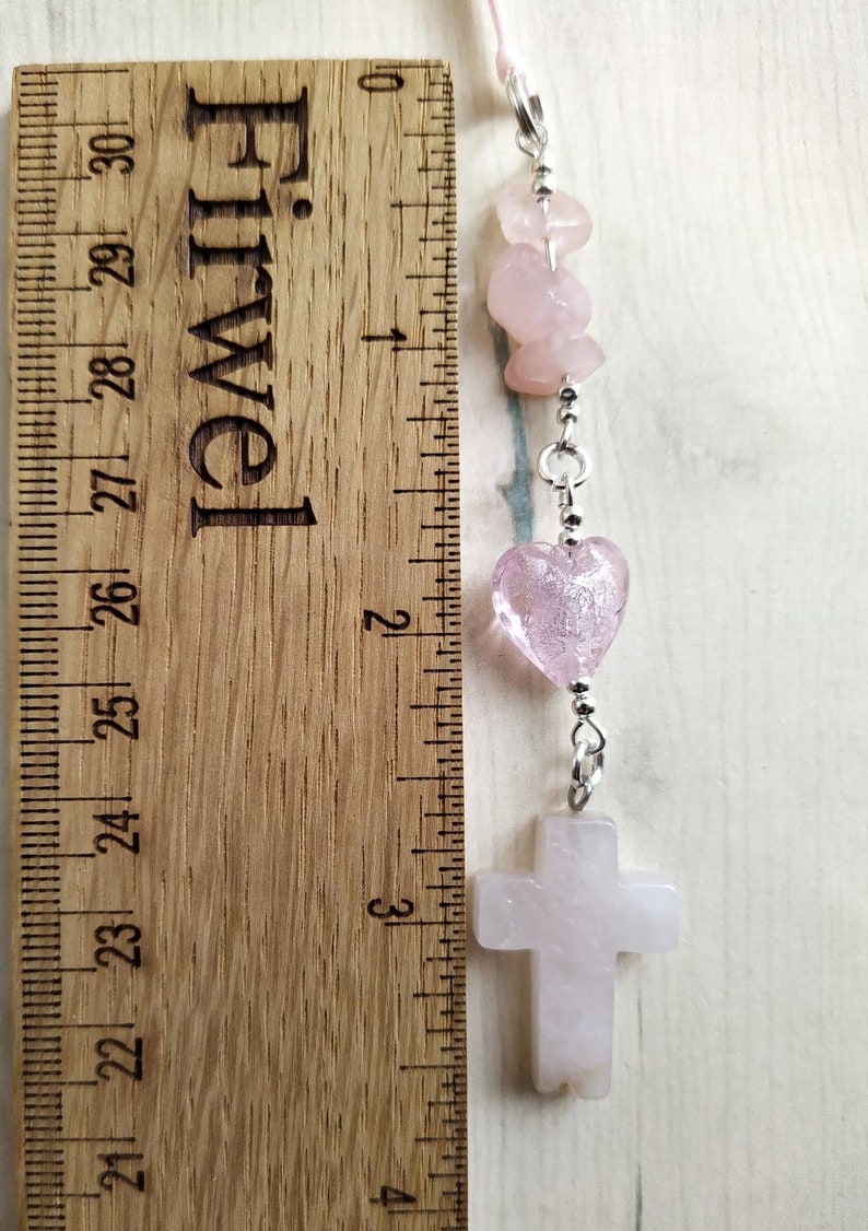 Puede incluir: Un brazalete de charms rosa y plateado con una cruz, corazones y cuentas. El brazalete mide aproximadamente 15 cm de largo.