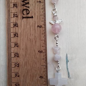Puede incluir: Un amuleto de cristal rosa y blanco con un amuleto de &aacute;ngel plateado y un amuleto de cruz plateado. El amuleto est&aacute; unido a una cinta rosa y se mide con una regla.