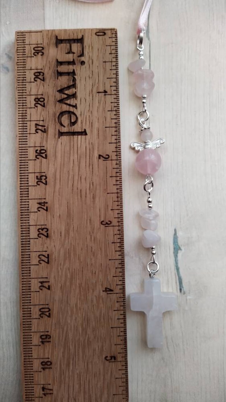 Puede incluir: Un amuleto de cristal rosa y blanco con un amuleto de &aacute;ngel plateado y un amuleto de cruz plateado. El amuleto est&aacute; unido a una cinta rosa y se mide con una regla.