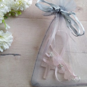 Puede incluir: Un dije de cruz rosa con detalles plateados est&aacute; empaquetado en una bolsa de organza gris transparente con una cinta plateada.