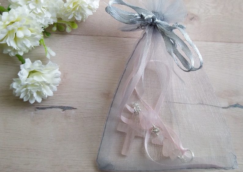 Puede incluir: Un dije de cruz rosa con detalles plateados est&aacute; empaquetado en una bolsa de organza gris transparente con una cinta plateada.