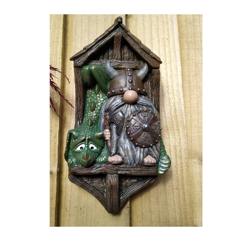 K&ouml;nnte beinhalten: Dekorative Wandplakette mit einem Wikinger-Gnom und einem gr&uuml;nen Drachen. Der Gnom tr&auml;gt einen geh&ouml;rnten Helm und h&auml;lt einen Schild und einen Speer. Der Drache hat einen traurigen Ausdruck. Die Plakette ist braun und gr&uuml;n.