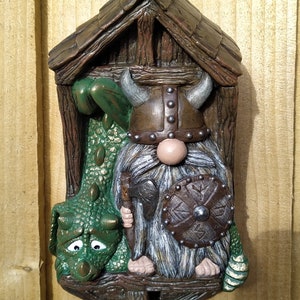 K&ouml;nnte beinhalten: Ein skurriles Wandbild mit einem Wikinger-Zwerg und einem gr&uuml;nen Drachen. Der Zwerg tr&auml;gt einen geh&ouml;rnten Helm und h&auml;lt Schild und Axt. Der Drache hat einen traurigen Gesichtsausdruck. Das St&uuml;ck wird von einer Holzkonstruktion umrahmt.