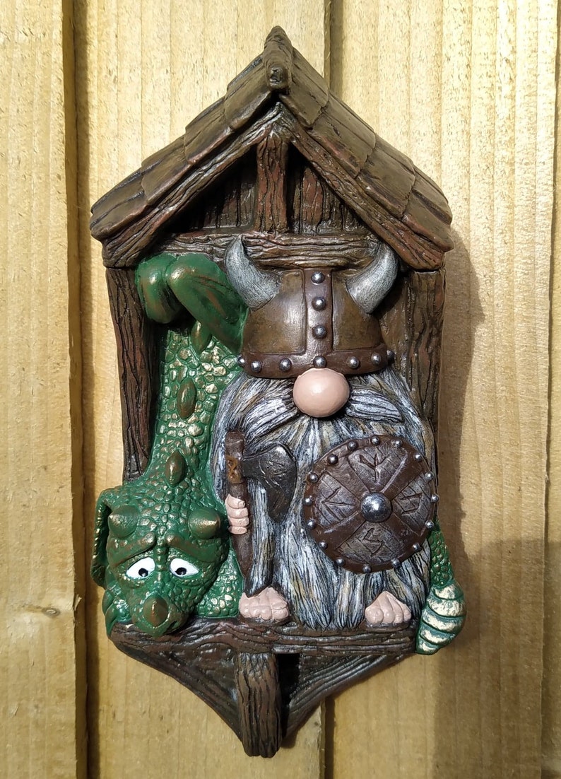 K&ouml;nnte beinhalten: Ein skurriles Wandbild mit einem Wikinger-Zwerg und einem gr&uuml;nen Drachen. Der Zwerg tr&auml;gt einen geh&ouml;rnten Helm und h&auml;lt Schild und Axt. Der Drache hat einen traurigen Gesichtsausdruck. Das St&uuml;ck wird von einer Holzkonstruktion umrahmt.