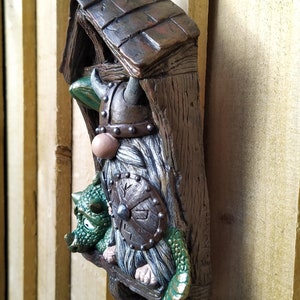 K&ouml;nnte beinhalten: Ein skurriles Wandbild mit einem Wikinger-Zwerg und einem gr&uuml;nen Drachen. Der Zwerg tr&auml;gt einen geh&ouml;rnten Helm und h&auml;lt einen Schild. Das St&uuml;ck ist aus Kunstharz gefertigt und hat ein verwittertes, holz&auml;hnliches Aussehen.