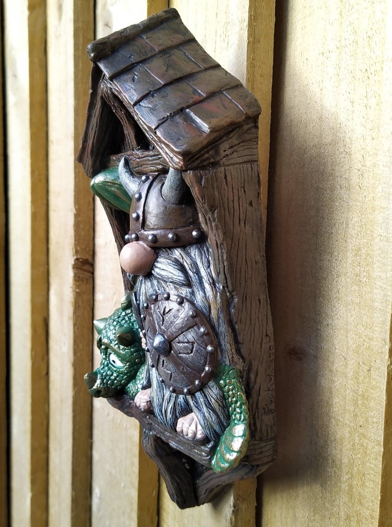 K&ouml;nnte beinhalten: Ein skurriles Wandbild mit einem Wikinger-Zwerg und einem gr&uuml;nen Drachen. Der Zwerg tr&auml;gt einen geh&ouml;rnten Helm und h&auml;lt einen Schild. Das St&uuml;ck ist aus Kunstharz gefertigt und hat ein verwittertes, holz&auml;hnliches Aussehen.