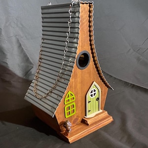 May include: A wooden birdhouse with a brown body, a gray corrugated metal roof, and a green door and window. A small chain is attached to the roof for hanging. The birdhouse has a whimsical, fairy-tale design.