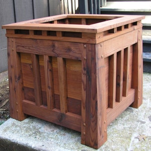 May include: A square wooden planter box with a dark brown stain. The planter has a decorative top with a slatted design and a raised edge. The planter is made of solid wood with visible grain.