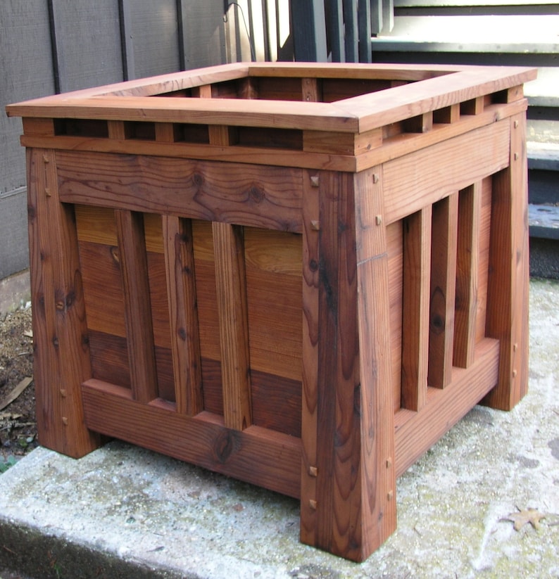 May include: A square wooden planter box with a dark brown stain. The planter has a decorative top with a slatted design and a raised edge. The planter is made of solid wood with visible grain.
