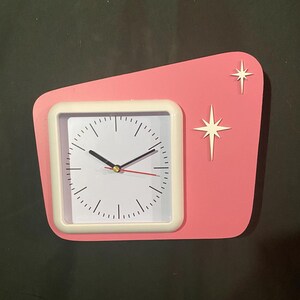 May include: A retro-style pink wall clock with a white square clock face and a white frame. Two white starbursts are on the pink background. The clock hands are black, with a red second hand.