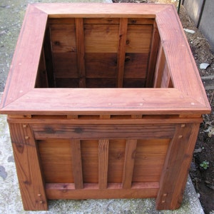 May include: A wooden planter box with a square shape and a raised rim. The planter is made of natural wood with a reddish-brown color. The planter has a simple design with a grid pattern on the front and sides.