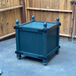 May include: A dark teal planter with black metal accents and decorative finials. The planter is made of wood and has a square shape.