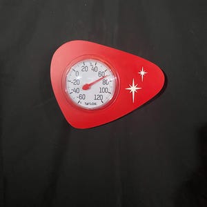 May include: A red, retro-style thermometer with a white face and black numbers. The thermometer reads in Fahrenheit, with a range from -60 to 120 degrees. The red casing has a unique shape with two white starburst designs.