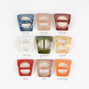 Op de afbeelding: Negen keramische toggle-sluitingen in verschillende kleuren, waaronder blauw, wit, groen, zand, rood, bruin, oranje, cacao en wijn. Elke sluiting heeft een uniek textuur.