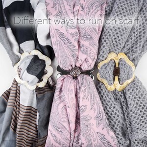 May include: Three different scarves with decorative scarf rings. The scarves are a black and white striped scarf, a pink paisley scarf, and a grey knitted scarf. The scarf rings are a white flower-shaped ring, a silver ring with a rose design, and a white flower-shaped ring with a brown button.