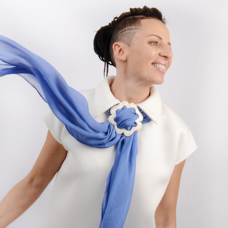 May include: A woman wearing a white collared shirt with a blue scarf tied around her neck. The scarf is secured with a white flower-shaped clasp.