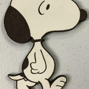 May include: A white wooden cutout of Snoopy, a cartoon beagle, walking with his signature black ear and nose. The cutout is a simple design with a brown outline.