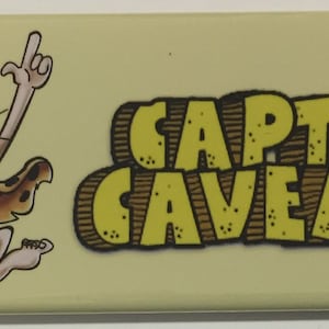 Puede incluir: Un imán amarillo y marrón con un personaje de dibujos animados con un garrote y el texto "CAPTAIN CAVEMAN".