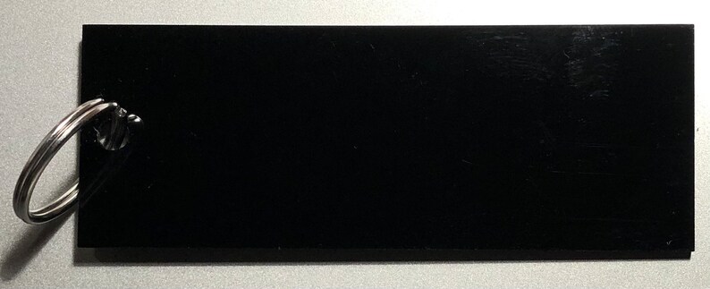 May include: A black rectangular tag with a silver key ring attached. The tag is blank and has a smooth surface.