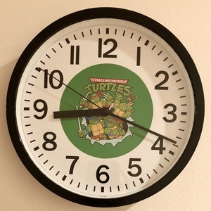 May include: Round wall clock with a black frame and white face. The clock features black numerals and hands, with a green circle in the center displaying the Teenage Mutant Ninja Turtles logo and characters.