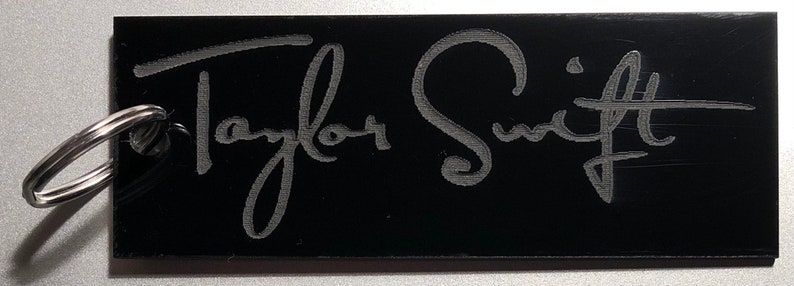 May include: Black keychain with a silver key ring and the engraved text "Taylor Swift" in a cursive font.