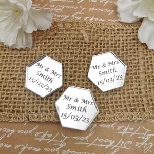 May include: Three silver hexagonal mirror tiles with the text "Mr & Mrs Smith 15/03/23" engraved on each. The tiles are arranged on a brown burlap surface.