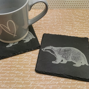 Może przedstawiać: Dwa czarne podkładki ze łupka z białym wzorem borsuka na jednej i białym wzorem serca na drugiej. Szary ceramiczny kubek z białym wzorem serca stoi na podkładce z wzorem serca.