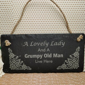 May include: A black slate sign with a white rope hanger. The sign has white floral designs on the sides and the text "A Lovely Lady And A Grumpy Old Man Live Here" is engraved in white.