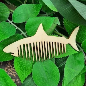 Puede incluir: Un peine de madera con forma de aleta de tiburón, con un acabado de madera natural. El peine está sobre hojas verdes.