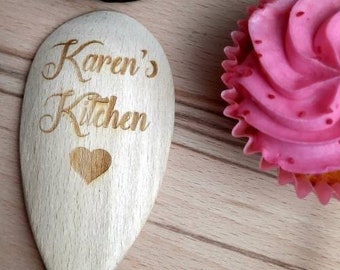 May include: A wooden spoon with the text "Karen's Kitchen" and a heart engraved on it. The spoon is surrounded by cupcakes and baking supplies.