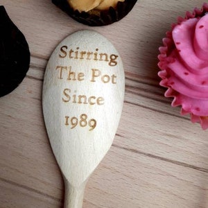May include: A wooden spoon with the text "Stirring The Pot Since 1989" engraved on it. The spoon is surrounded by cupcakes and baking supplies.