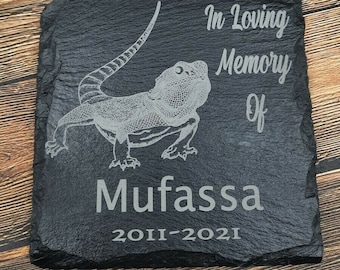 May include: Black slate memorial plaque with a white engraved image of a bearded dragon. The text reads "In loving memory of Mufassa 2011-2021".