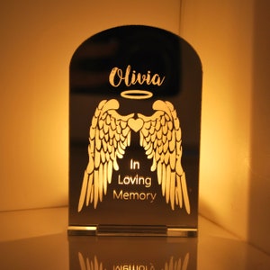 May include: A mirrored memorial candle holder with a halo and angel wings design. The text "Olivia" and "In Loving Memory" is engraved on the holder.