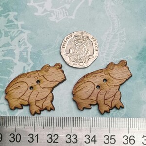 Peut inclure: Deux boutons en bois en forme de grenouille avec une finition en bois brun. Les boutons mesurent environ 2,5 cm de diamètre et présentent un motif de grenouille détaillé. Les boutons sont présentés à côté d'une règle et d'une pièce de 20 pence.
