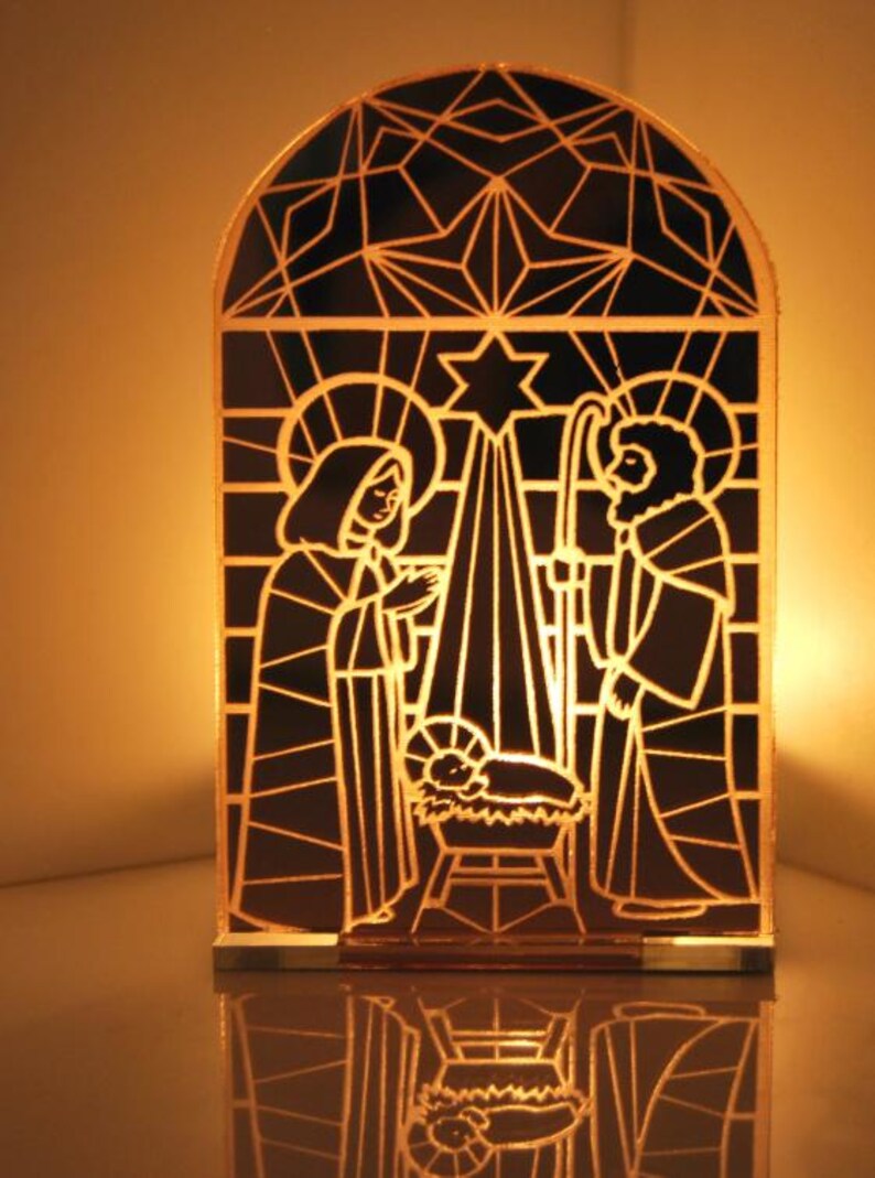 Puede incluir: Una escena de la Natividad de Navidad de estilo vitral con Mar&iacute;a, Jos&eacute; y el ni&ntilde;o Jes&uacute;s en un pesebre. La escena est&aacute; iluminada desde atr&aacute;s, creando un brillo c&aacute;lido.