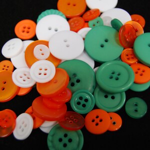 Puede incluir: Una colección de botones de varios tamaños y colores, incluyendo naranja, blanco y verde. Los botones, con dos o cuatro agujeros, están dispersos sobre un fondo negro. Algunos son redondos, otros ovalados.