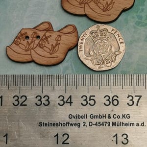May include: Two pairs of wooden buttons shaped like Dutch clogs with a windmill and tulip design. The buttons are approximately 1.5 inches in diameter.  The buttons are shown next to a 20 pence coin for size reference.