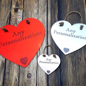 Può includere: Tre cartelli di legno a forma di cuore con ganci in corda. I cartelli sono rossi, bianchi e in legno naturale. Ogni cartello ha il testo "Any Personalisation" stampato su di esso.