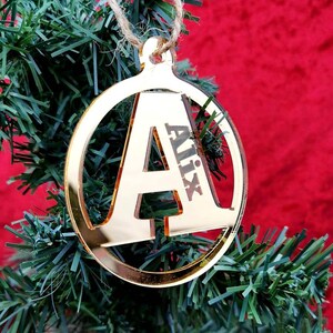 Puede incluir: Adorno de espejo dorado con la letra A y el nombre Alix inscrito en él. El adorno está colgado de un árbol de Navidad artificial verde.