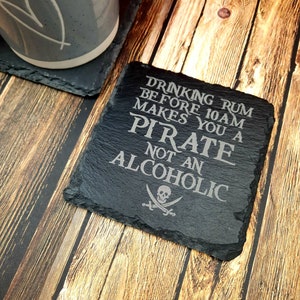 May include: A black slate coaster with the engraved text "DRINKING RUM BEFORE 10AM MAKES YOU A PIRATE NOT AN ALCOHOLIC" and a skull and crossbones symbol. A gray mug with a heart design is visible in the background.
