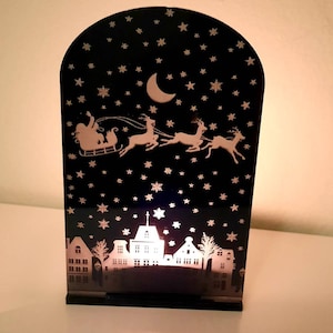 Puede incluir: Una luz decorativa negra con temática navideña con una silueta de Papá Noel en un trineo tirado por renos bajo un cielo estrellado. La parte inferior presenta una silueta de ciudad. La luz está sobre una superficie blanca.