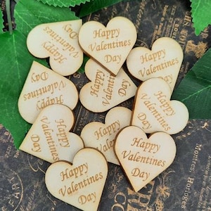 Op de afbeelding: Een verzameling houten hartvormige ornamenten met de tekst "Happy Valentines Day" gegraveerd op elk stuk. De ornamenten zijn verspreid over een donkere achtergrond met een goud-zwart patroon.