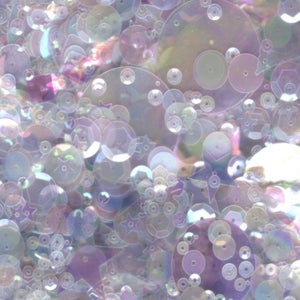 May include: A collection of iridescent sequins in various shapes and sizes, including circles and stars. The sequins are a mix of light purple, white, and clear colors.