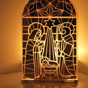 Puede incluir: Una escena de la Natividad de Navidad de estilo vitral con Mar&iacute;a, Jos&eacute; y el ni&ntilde;o Jes&uacute;s en un pesebre. La escena est&aacute; iluminada desde atr&aacute;s, creando un brillo c&aacute;lido.